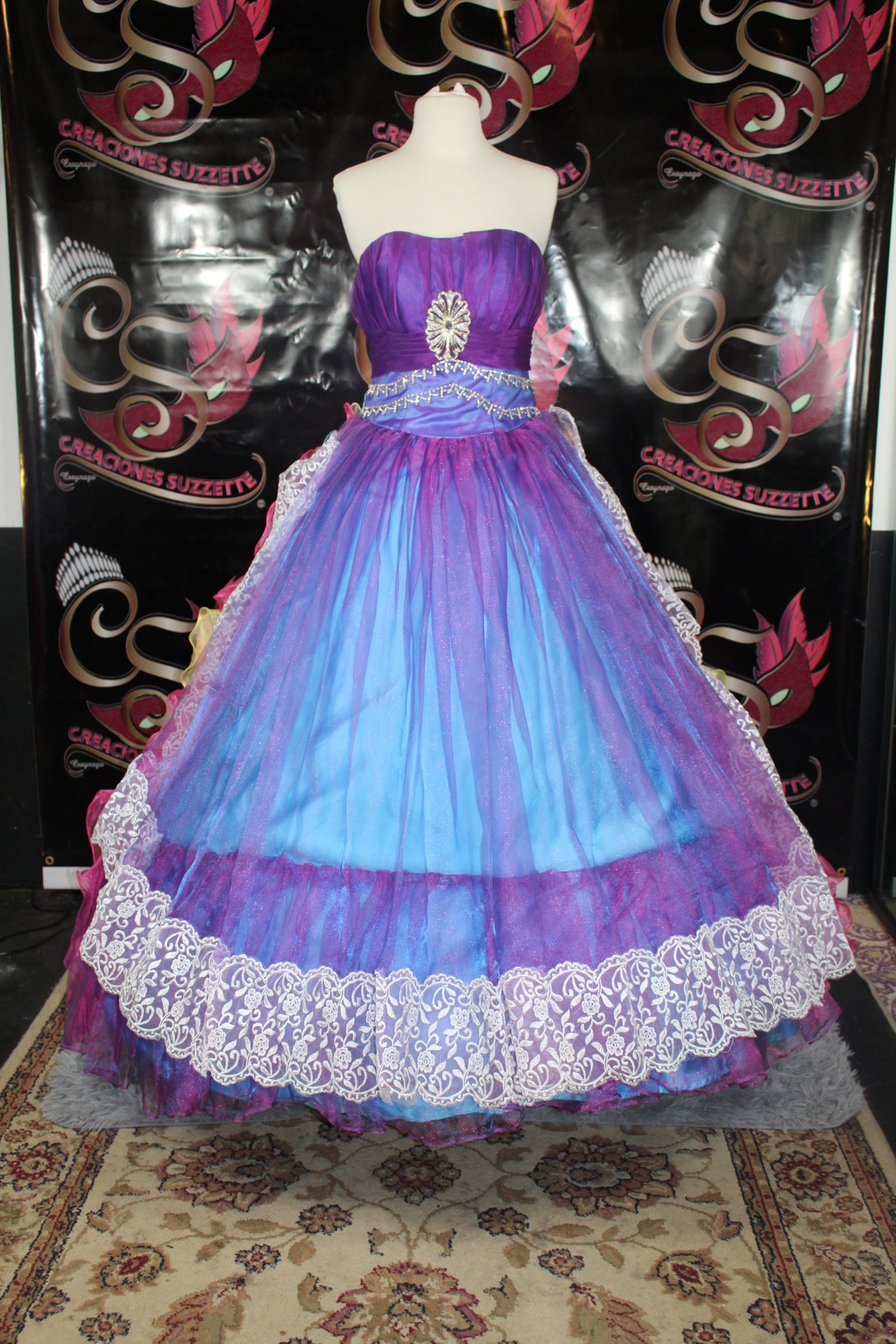 A strapless ball gown displayed on a mannequin, featuring a purple bodice with a silver jewel detail, a blue and purple gradient waist sash, and a voluminous tulle skirt in blue, purple, and pink with a white lace hem. The background is a black printed banner with gold and pink masks.