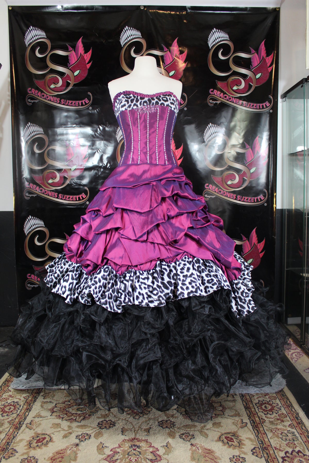 A voluminous gothic-style ball gown displayed on a mannequin torso in front of a black decorative backdrop. The dress features a purple and magenta corset bodice with silver trim, layered ruffles in purple and black, leopard-print midriffs, and a large black ruffled tulle skirt.