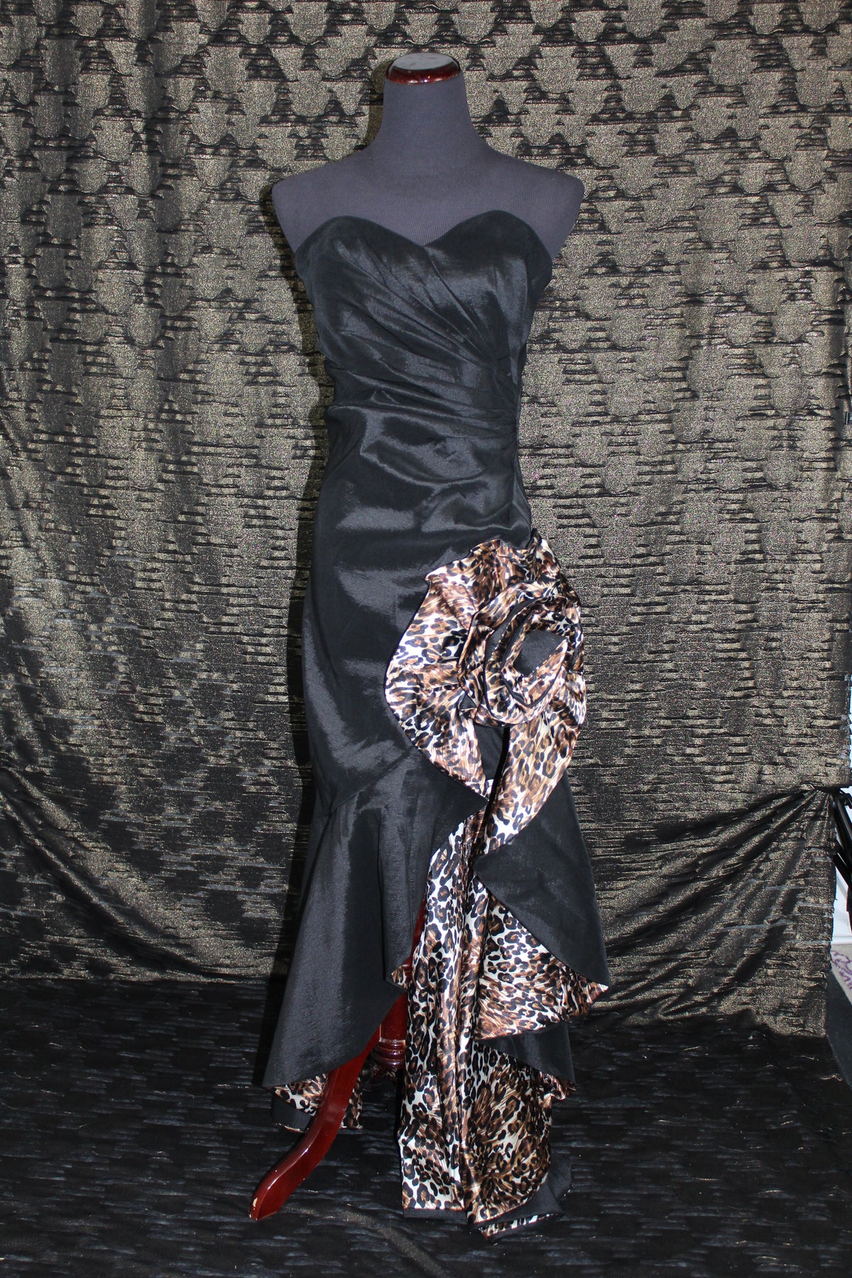 A strapless black satin gown displayed on a mannequin, featuring a large draped floral bow in a leopard print pattern on the skirt, set against a patterned gray fabric backdrop.