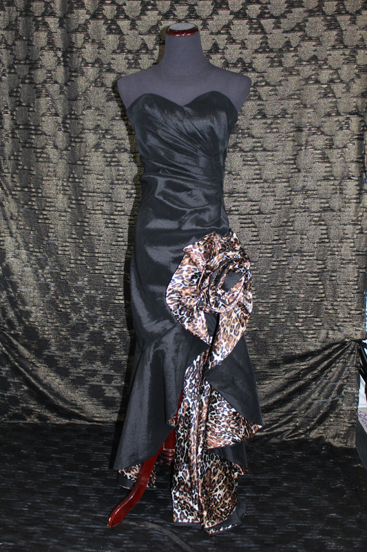 A strapless black satin gown displayed on a mannequin, featuring a large draped floral bow in a leopard print pattern on the skirt, set against a patterned gray fabric backdrop.