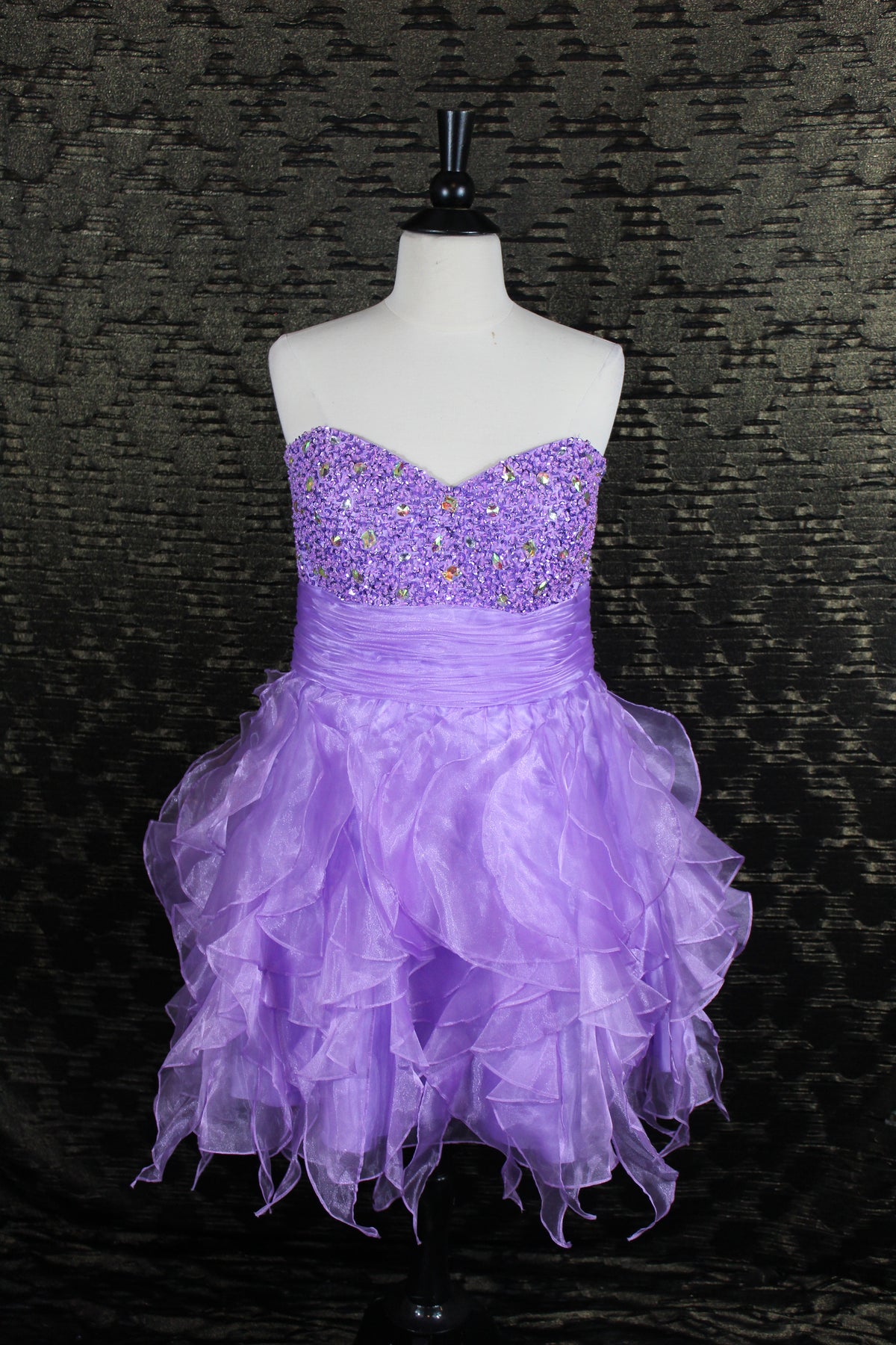 A lavender purple strapless tulle dress with a beaded sequin sweetheart bodice, displayed on a white mannequin torso against a dark textured backdrop.