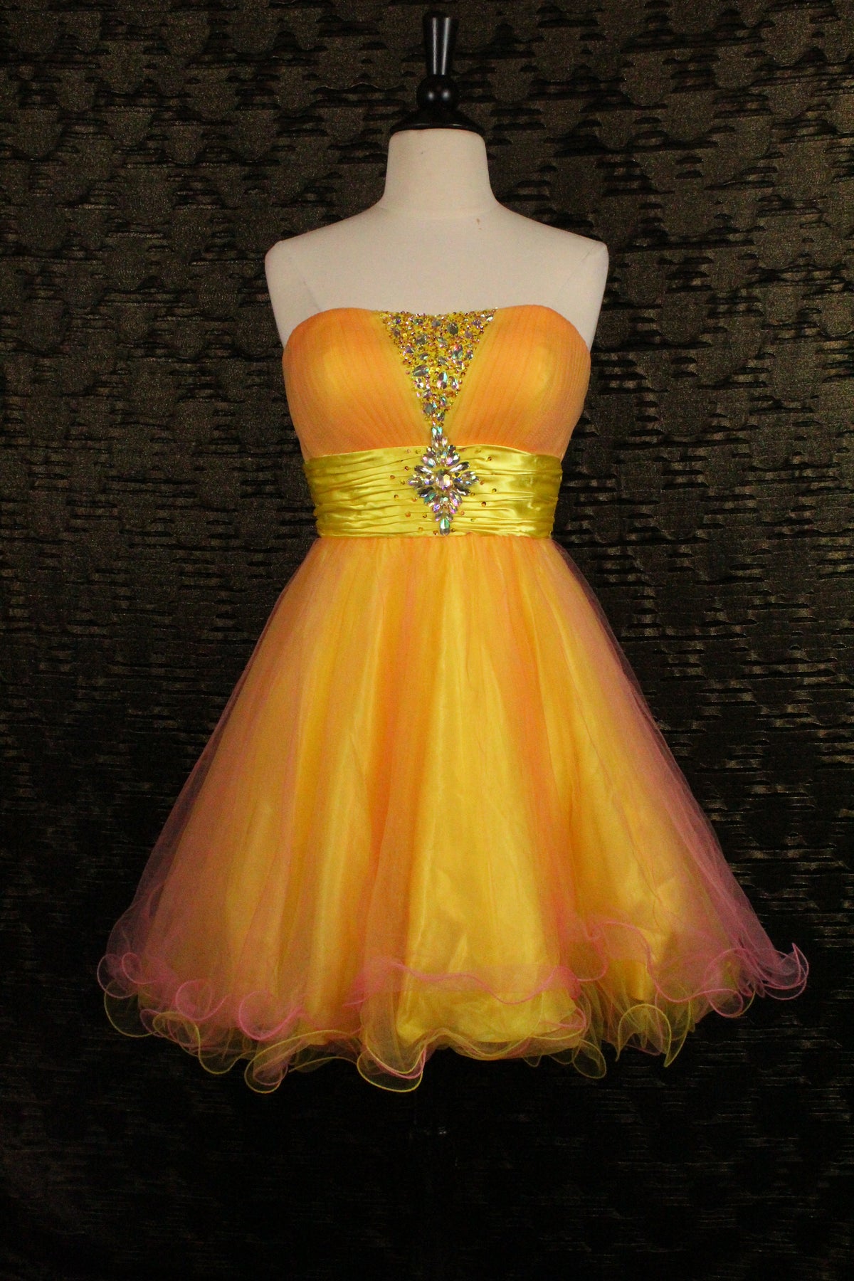 A strapless yellow and orange tulle homecoming dress displayed on a white mannequin torso against a dark textured background. The dress features a yellow satin waistband with a jeweled embellishment at the center bust.
